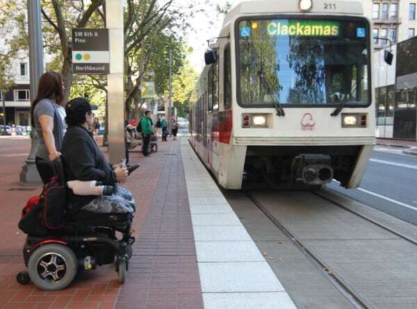 A TriMet light rail pulls up as an MTM Transit assessment evaluator and a man who utilizes a wheelchair looks on. MTM Transit helps TriMet ensure LIFT paratransit eligibility.