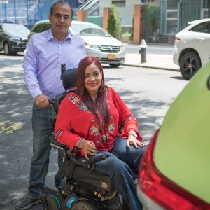 A driver stands behind a woman who utilizes a wheelchair. On behalf of the TLC's Accessible Dispatch program, MTM dispatches accessible taxis to New Yorkers with disabilities.