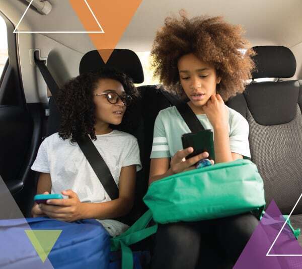 Two young girls look at their phones while sitting in a vehicle. MTM Transit offers reliable student transportation and special needs transportation to schools and programs serving children, including those covered by the McKinney-Vento Act.